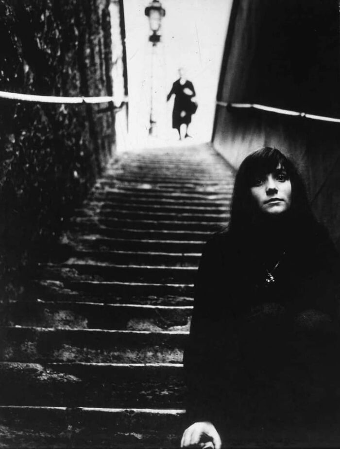 Black and white vintage photo of a woman sitting on stone stairs with a blurred figure descending in the background.