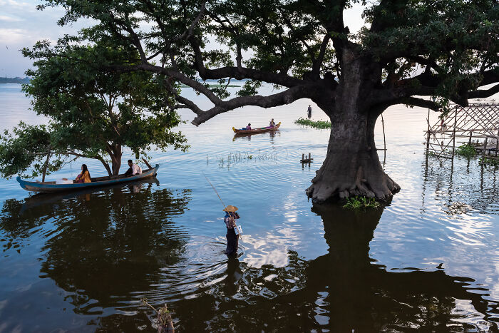 Fisherman and boats near large trees in a calm lake, capturing breathtaking travel photography award-winning scenery.