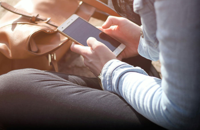 Person holding smartphone reading unhinged and disturbing voicemails outdoors with brown leather bag nearby in daylight.