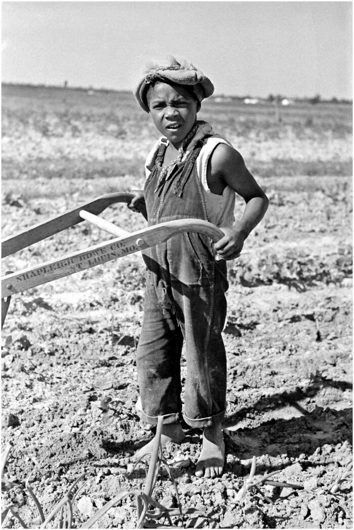 Young boy working barefoot in a field using a plow, historical vintage photo showcasing early farming practices.