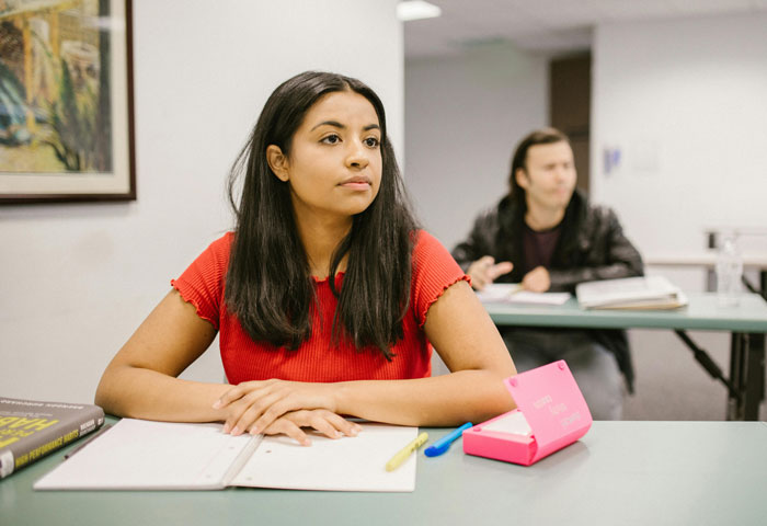 Young woman in red shirt sitting at desk with notebook and pink phone, appearing distracted by disturbing voicemails received.