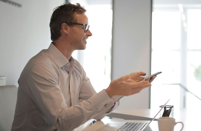 Man in office wear holding a phone gesturing while working at a laptop, illustrating jobs done by awful people concept.