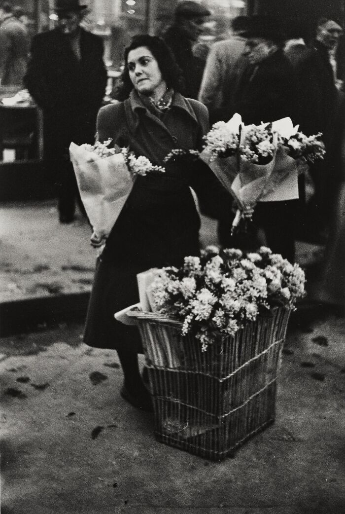 Woman holding bouquets of flowers and standing by a basket of vintage flowers in a historical black and white photo.