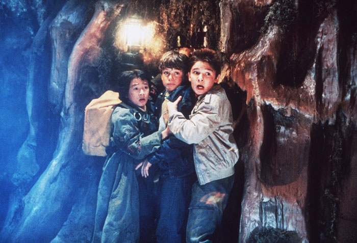 A tense scene from the movie The Goonies featuring three young boys huddled together in a narrow cave passage, looking frightened and alert. A lantern glows behind them, casting dim light on the rugged, damp walls. The boys wear jackets and backpacks, appearing lost and nervous as they brace for something unknown. A tense scene from the movie The Goonies featuring three young boys huddled together in a narrow cave passage, looking frightened and alert. A lantern glows behind them, casting dim light on the rugged, damp walls. The boys wear jackets and backpacks, appearing lost and nervous as they brace for something unknown.