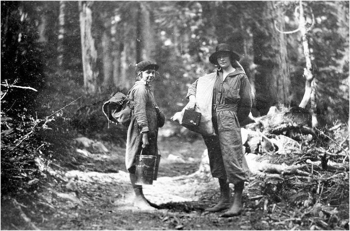 Two women dressed in early 20th-century attire carrying supplies, featured in a historical and vintage photos collection.