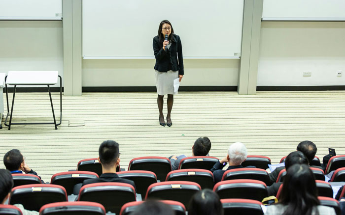 Woman giving a presentation in a lecture hall to an audience, illustrating jobs once respected but done by awful people.