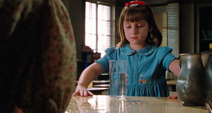 A scene from the movie Matilda featuring a young girl with a red ribbon in her hair and wearing a blue dress, concentrating intensely while staring at a glass of water on a table. The water has spilled, and her hands are positioned near the glass as if she is trying to control it with her mind. The room is softly lit by sunlight coming through the blinds in the background. A scene from the movie Matilda featuring a young girl with a red ribbon in her hair and wearing a blue dress, concentrating intensely while staring at a glass of water on a table. The water has spilled, and her hands are positioned near the glass as if she is trying to control it with her mind. The room is softly lit by sunlight coming through the blinds in the background.