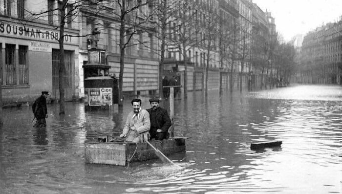 Two men navigating a flooded historical city street in a small wooden boat vintage photo from a historical photo collection.