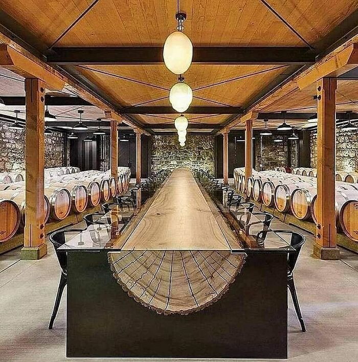 Large wood dining table made from a single slab in a rustic winery room with wooden beams and wine barrels.