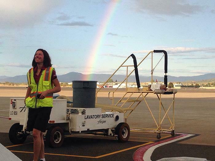 Airport worker smiling near lavatory service cart with a rainbow in the background, creating a confusing perspective.