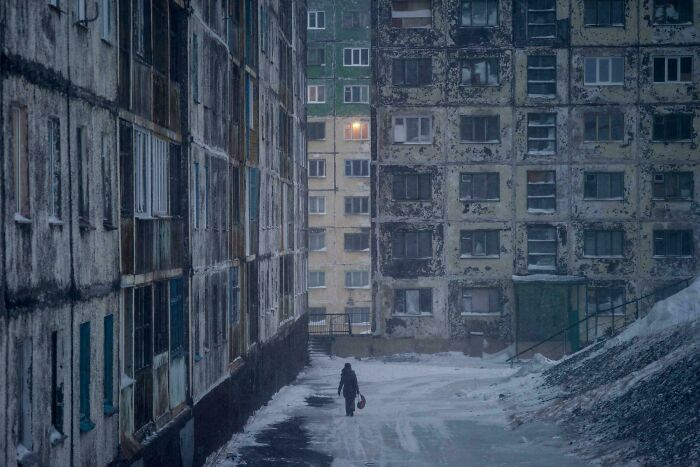 Urban hell scene with a solitary figure walking between dilapidated buildings in snowy weather.