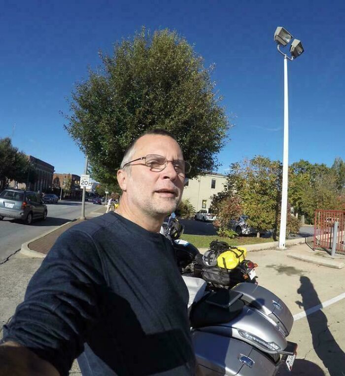 A man stands by a motorcycle with a tree aligned behind him, creating a confusing visual effect.