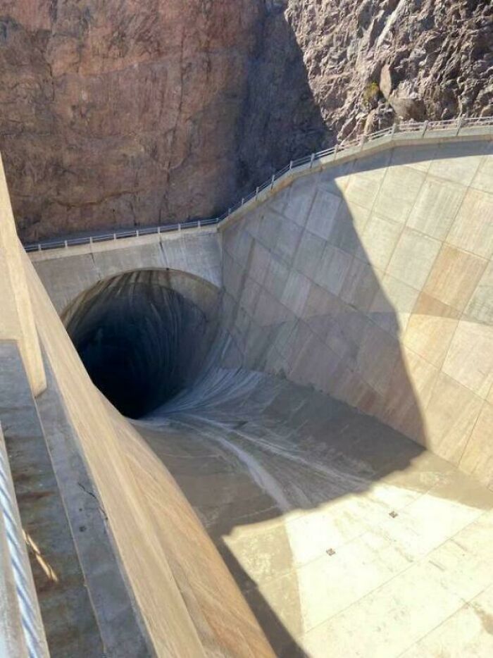 Massive concrete dam with a steep drop and large dark tunnel opening causing a sense of megalophobia fear.