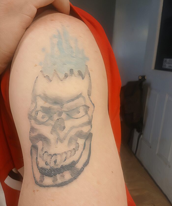 Tattoo of a distorted skull with blue flames on an arm, featured in a Facebook group for unusual tattoos.