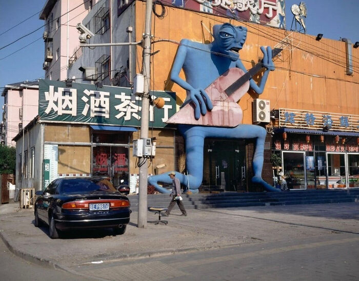 Blue bizarre building sculpture of a sitting creature playing a large guitar outside a commercial storefront in a city.