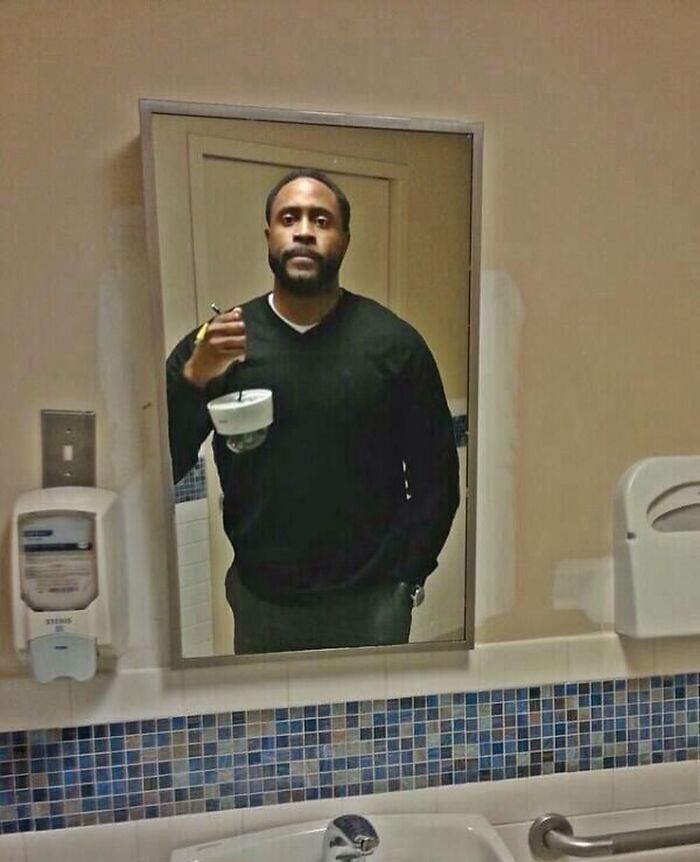Man in mirror holding coffee cup with toothbrush, wearing a black sweater in a tiled bathroom.