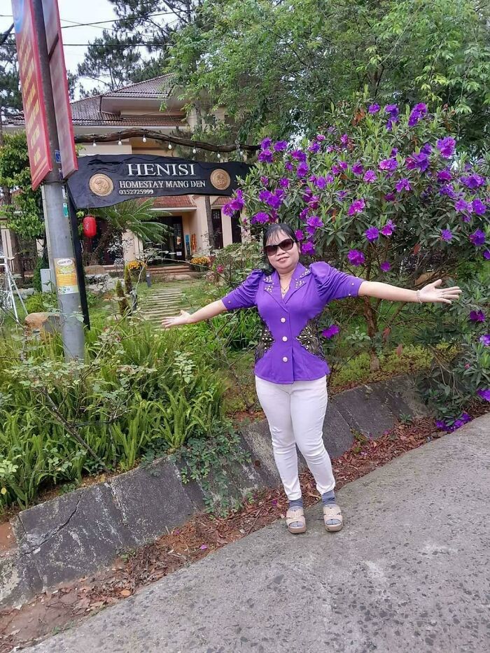 Woman in purple shirt posing in front of Henisi Homestay sign, surrounded by flowers, creating a confusing perspective.