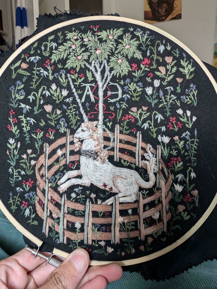 Creative embroidery featuring a unicorn within a circular fence, surrounded by colorful flowers on a black background.