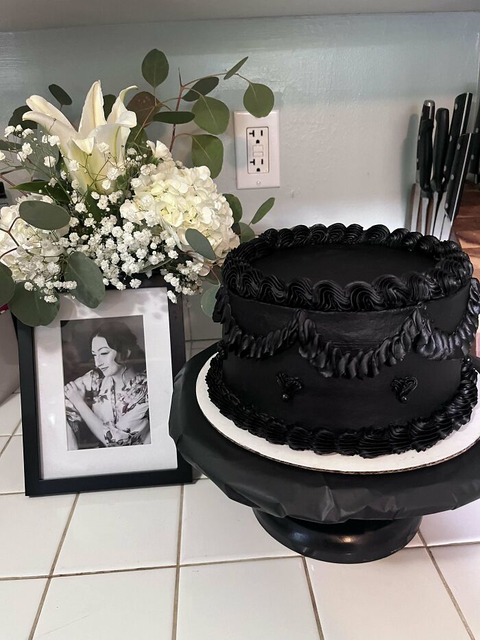 Black frosted cake with decorative piping on a stand next to white flowers and a framed black and white photo on a kitchen counter.