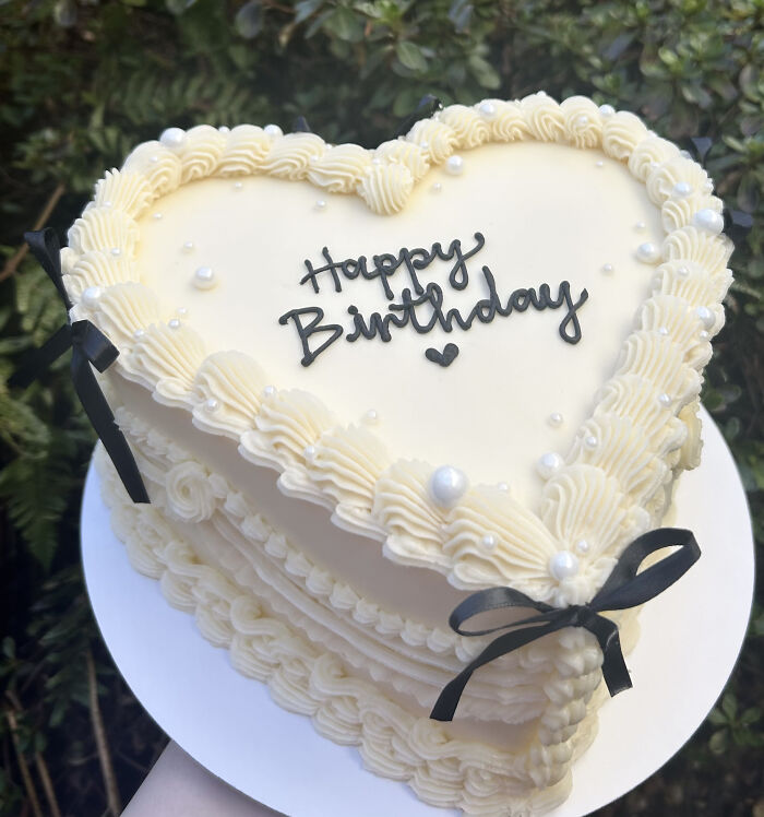 Heart-shaped birthday cake with white frosting, decorative pearls, and black ribbon, showcasing baking at its finest.