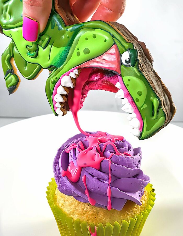 Hand holding a dinosaur-shaped cookie above a cupcake with purple frosting and pink icing drizzle in baking at its finest.