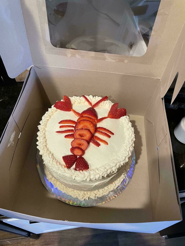Strawberry lobster-shaped decoration on a white frosted cake inside a bakery box, showcasing baking at its finest.