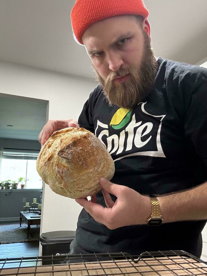 Man wearing an orange beanie holding a round loaf of homemade bread, showcasing baking at its finest.