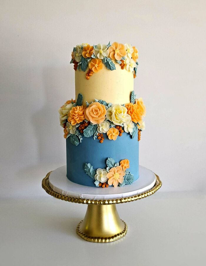 Two-tier floral cake with detailed buttercream flowers in yellow, orange, and blue, showcasing baking at its finest on gold stand.