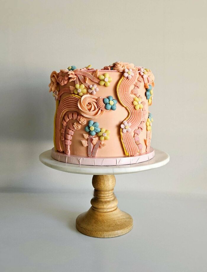 Decorative pink cake with floral and wavy piping details on a marble and wood cake stand, showcasing baking at its finest.