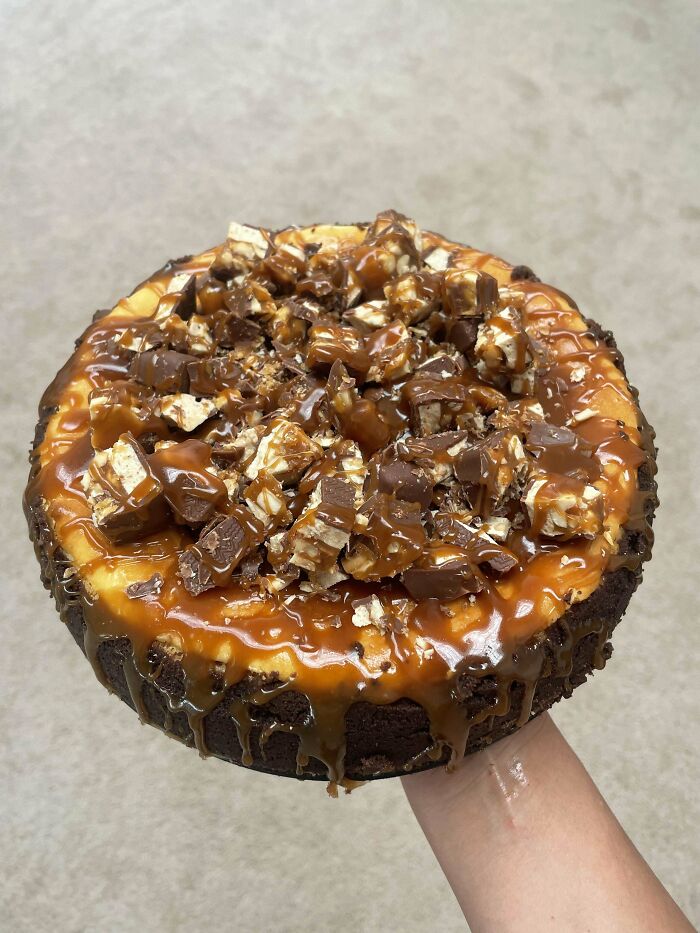 Hand holding a decadent cake topped with caramel drizzle and chopped chocolate pieces, showcasing baking at its finest.