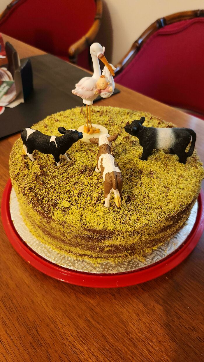 Round cake covered in crushed nuts featuring toy cows and a stork figurine, showcasing baking at its finest creativity.