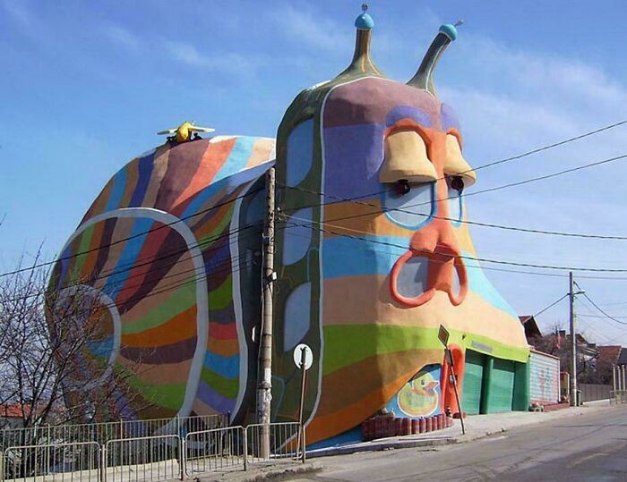 Colorful bizarre building shaped like a large snail with expressive eyes and a striped shell on a street corner.