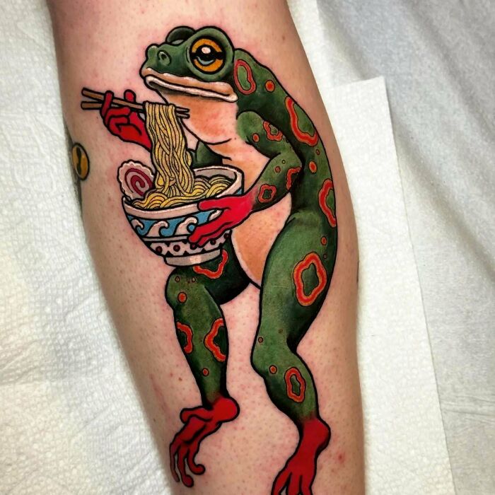 Tattoo of a colorful frog holding a bowl of noodles with chopsticks on someone's leg.