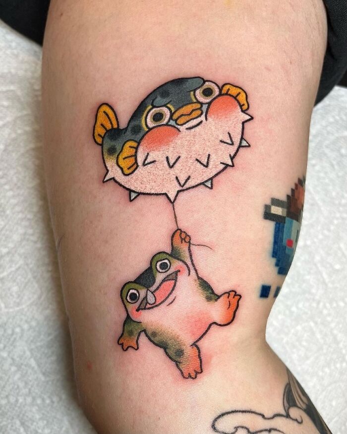 Colorful tattoo of a frog hanging from a pufferfish balloon on skin.