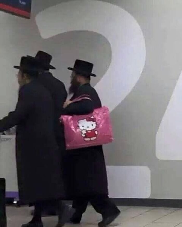 Men in black hats walking indoors with one holding a bright pink Hello Kitty bag, showcasing a humorous contrast.