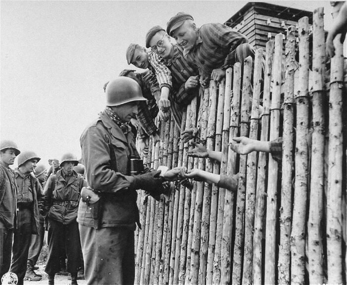 U.S. Army Corporal Larry Matinsk Puts Cigarettes Into The Extended Hands Of Newly Liberated Prisoners Behind A Stockade In The Allach Concentration Camp. April 30, 1945