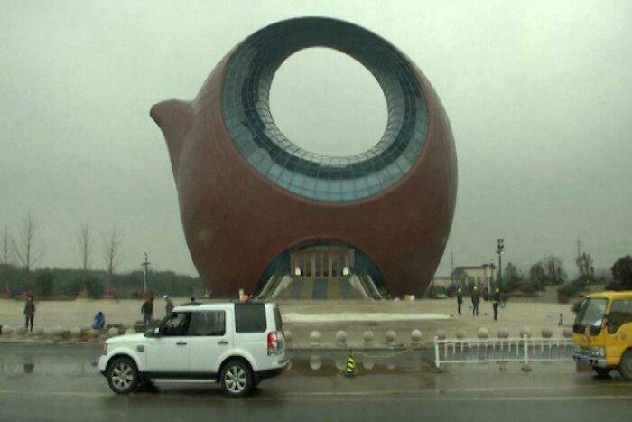 Unique bizarre building shaped like a giant teapot with a large circular window, surrounded by vehicles and people on a rainy day.