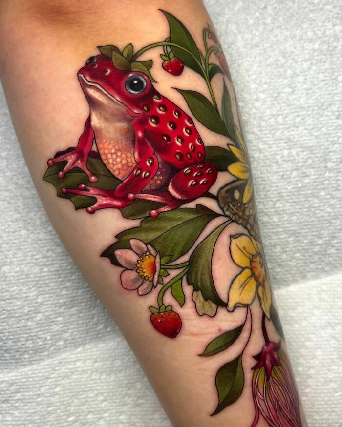 Colorful frog tattoo with floral and strawberry accents on an arm.