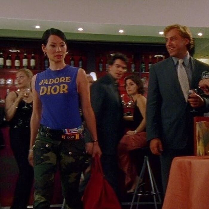 '90s fashion moment with a woman in a "J'adore Dior" shirt and camo pants at a lively bar scene.