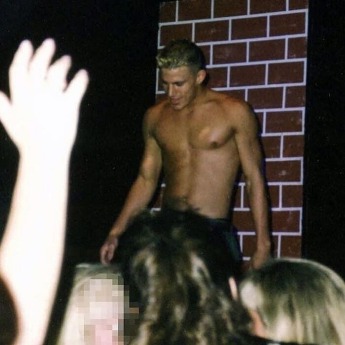 Shirtless man from the '90s standing by a brick wall, surrounded by a crowd with raised hands.