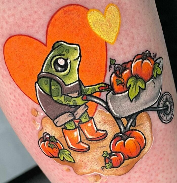 Frog tattoo with a frog pushing a pumpkin-filled wheelbarrow, set against orange heart shapes.