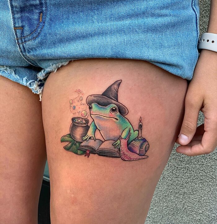 Frog tattoo on thigh depicting a frog with a wizard hat, cauldron, book, snail, and candle.