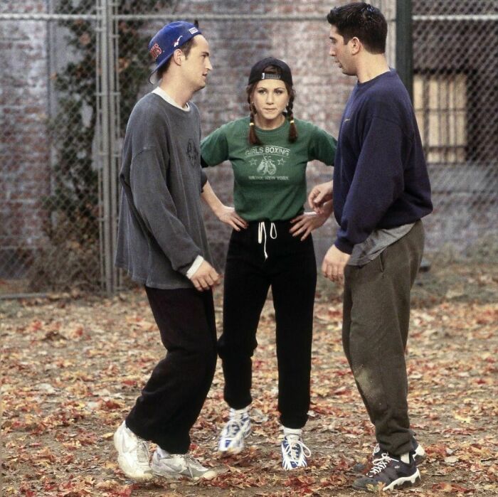 90s trio in casual outfits, standing outdoors on a leafy ground, chatting in an autumn setting.