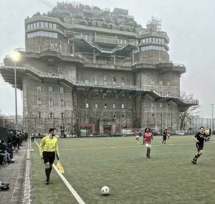 Large brutalist building with an ominous design overlooking a soccer field during a match, evoking supervillain headquarters vibes.