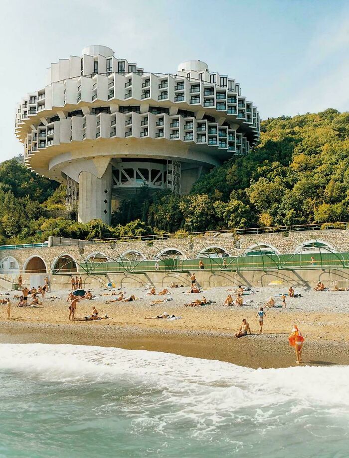 Unique bizarre building with futuristic design overlooking a beach crowded with sunbathers and waves crashing on shore.