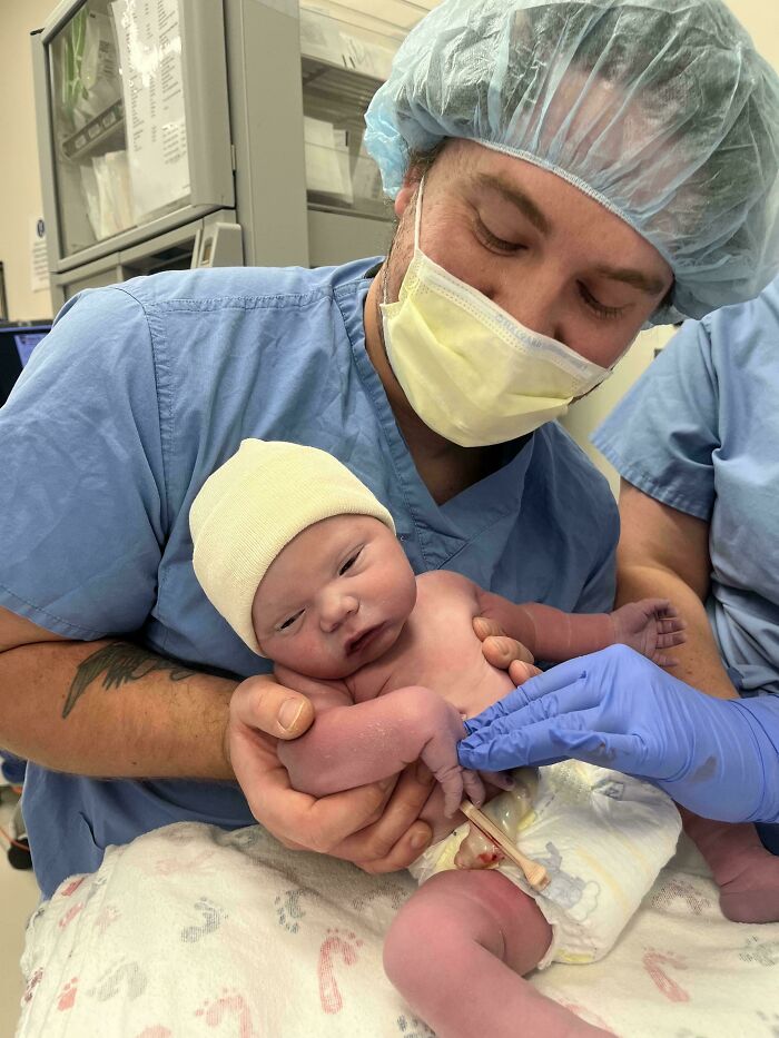 A wholesome dad in medical scrubs and mask gently holding his newborn baby moments after birth.