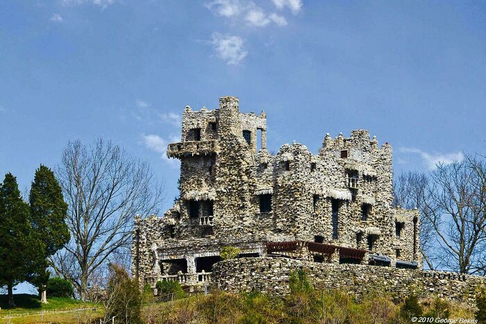 Bizarre building made of irregular stones resembling a castle, set against a clear blue sky and surrounding trees.
