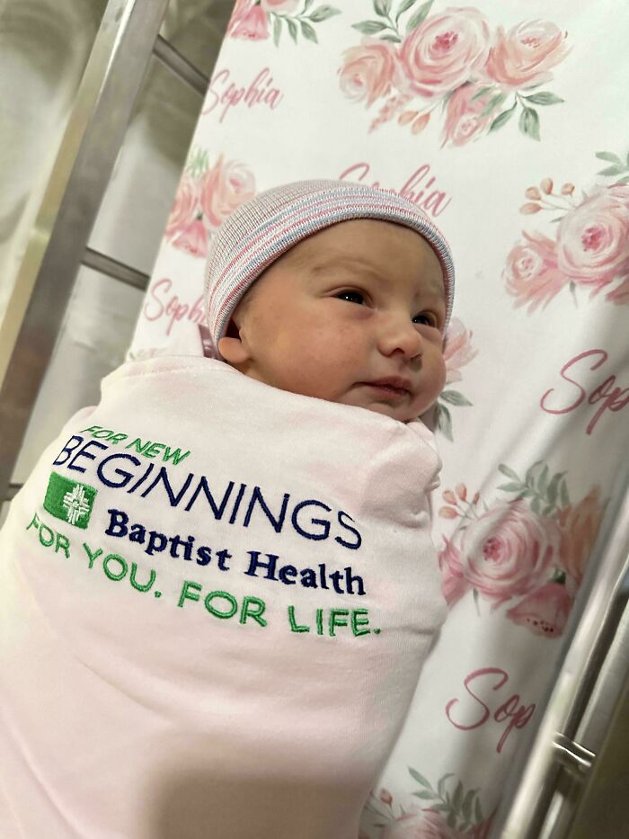 Bebé recién nacido envuelto en manta con logo Baptist Health en cuna con sábanas de flores y nombre Sophia.