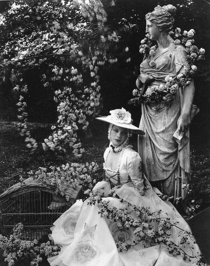Vintage photo of a woman in historical dress seated beside a floral-decorated statue, capturing a timeless vintage moment.