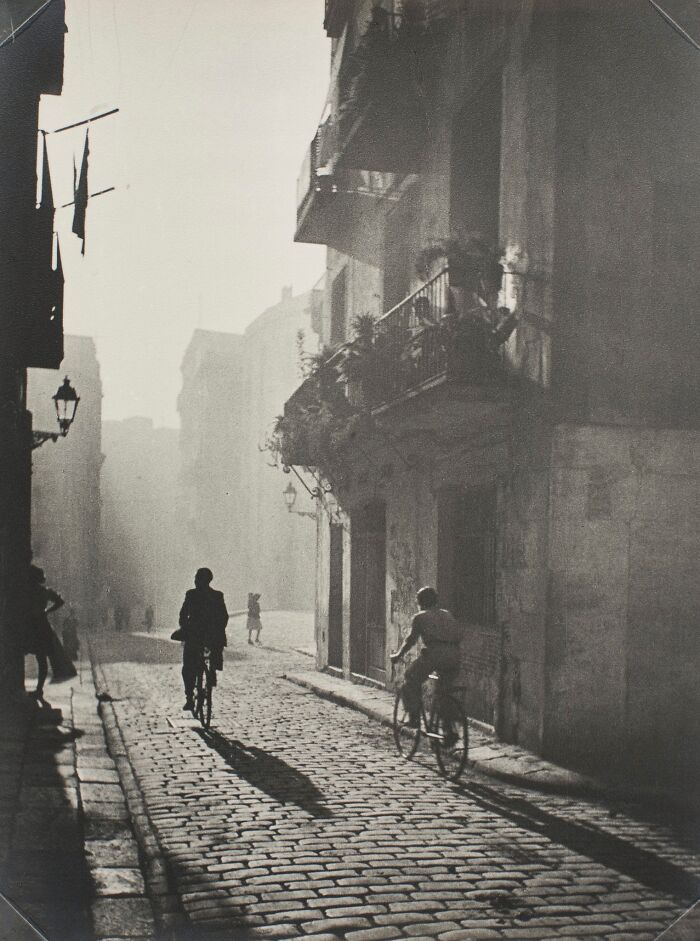 Vintage black and white photo showing people riding bicycles on a cobblestone street in a historical vintage setting.